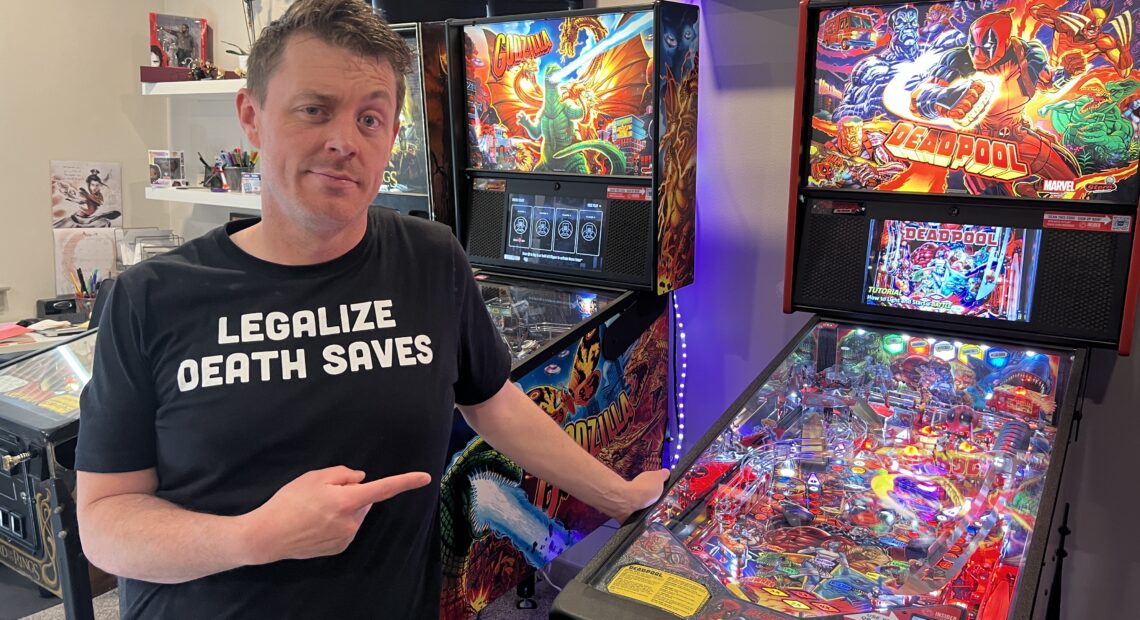 A man in a black shirt that says "legalize death saves" is standing in front of a multicolored Deadpool pinball machine.