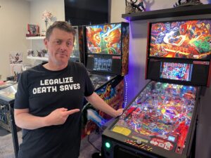 A man in a black shirt that says "legalize death saves" is standing in front of a multicolored Deadpool pinball machine.