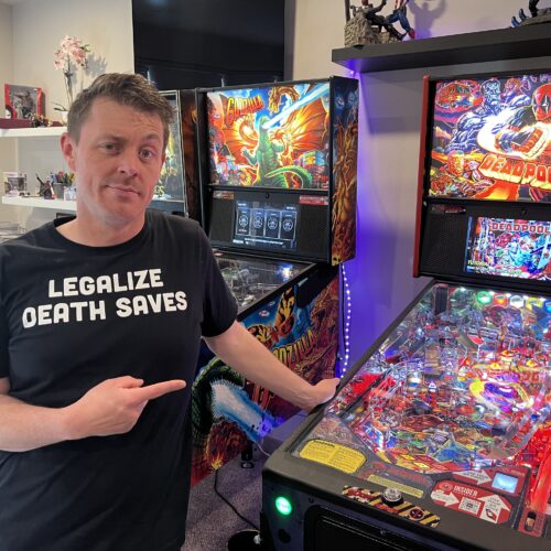 A man in a black shirt that says "legalize death saves" is standing in front of a multicolored Deadpool pinball machine.