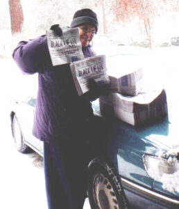 A woman in a beanie and purple coat holds up issues of a paper that reads "The Black Lens."