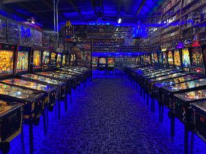 pinball machines line an aisle in an arcade the size of a warehouse. The room is lit in a bright blue light.