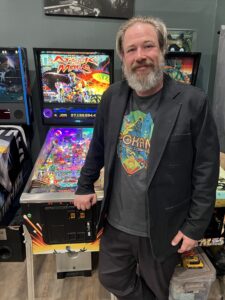 A man in a pinball T-shirt and a blazer stands in front of an Attack from Mars pinball machine.