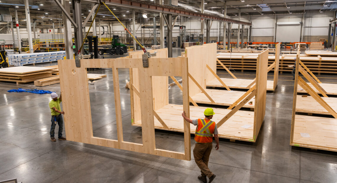 Workers in yellow hardhats and orange vests construct modular timber homes in a warehouse.