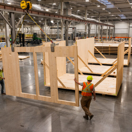 Workers in yellow hardhats and orange vests construct modular timber homes in a warehouse.