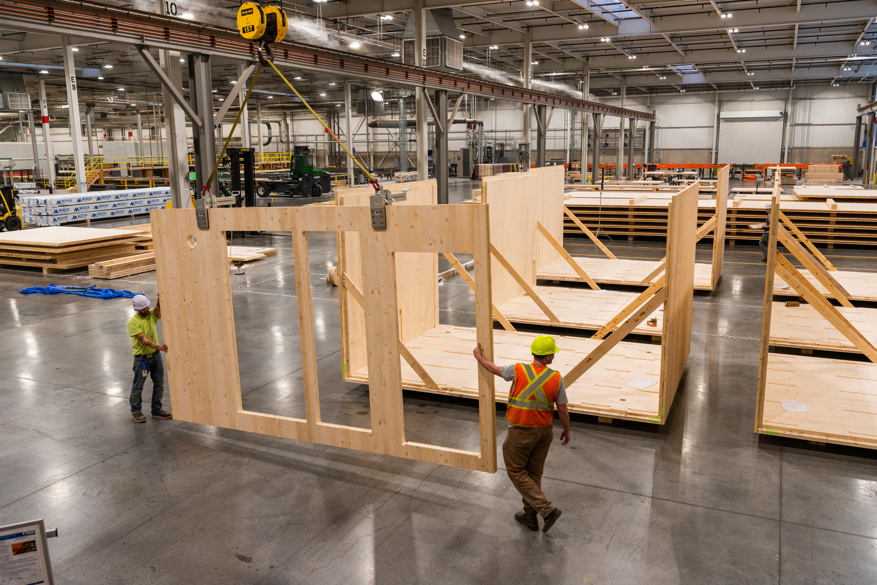 Workers in yellow hardhats and orange vests construct modular timber homes in a warehouse.