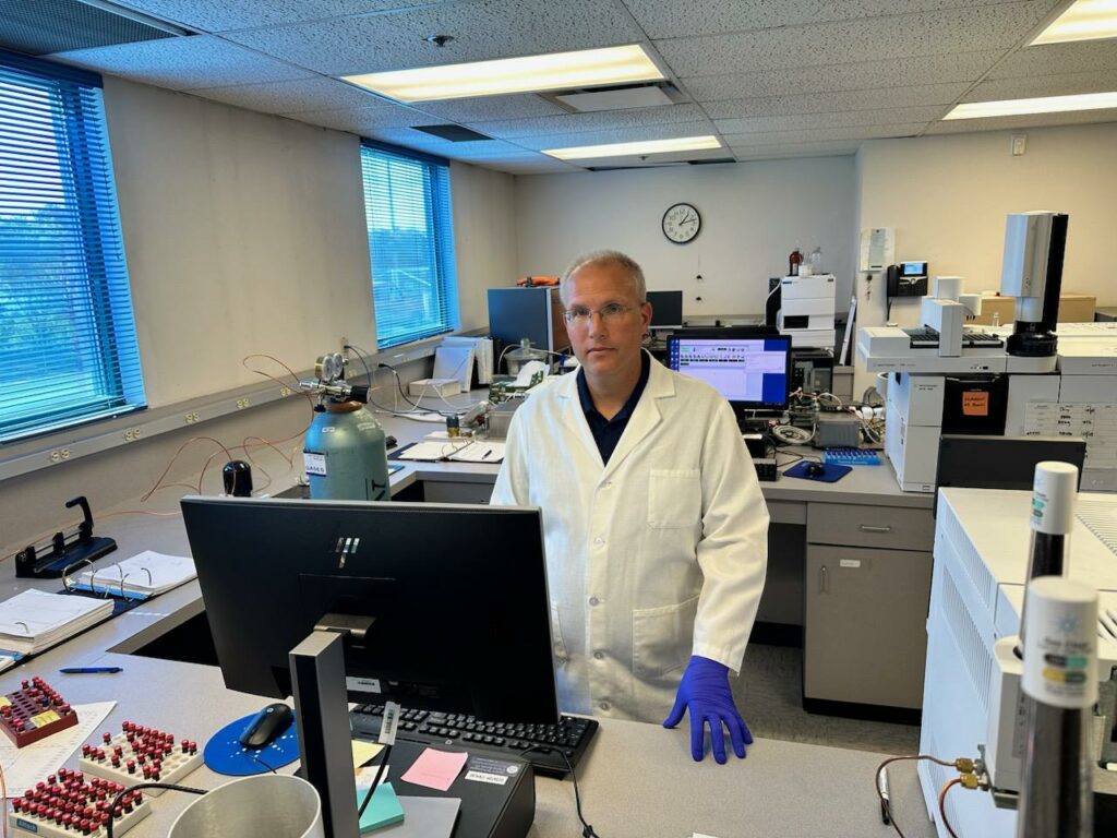 A man in a white labcoat with purple gloves and glasses on stands at his desk at the Idaho State Police Forensics Laboratory. 