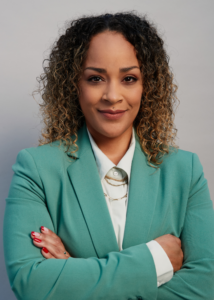 A woman with curly hair wears a teal blazer and smiles at the camera with her arms crossed.
