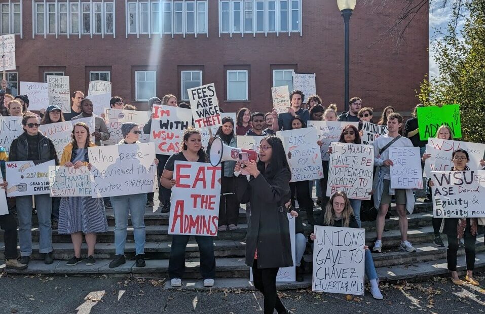 Students stand on steps holding signs with sayings like "Eat the Admins" in front of a brick building under a blue sky. A student gives a speech with a megaphone in front of the crowd.
