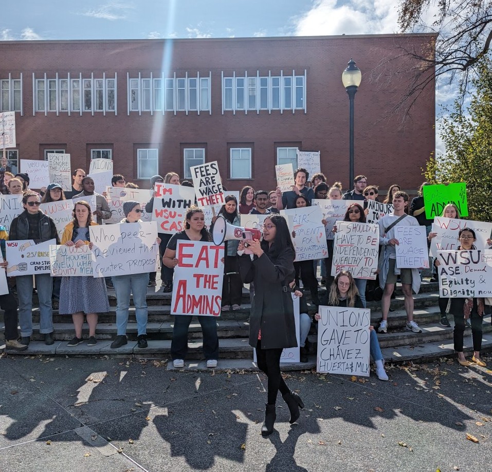 Students stand on steps holding signs with sayings like "Eat the Admins" in front of a brick building under a blue sky. A student gives a speech with a megaphone in front of the crowd.