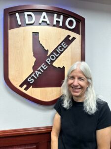 A woman with silver gray hair stands and smiles before a carved wood sign that reads "Idaho State Police." She is wearing a black t-shirt. 