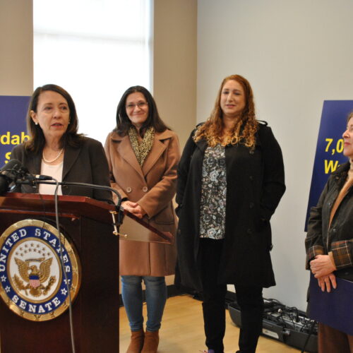 Senator Maria Cantwell stands at a brown podium alongside Spokane Mayor Lisa Brown, while two other affordable housing advocates in winter jackets stand behind them.
