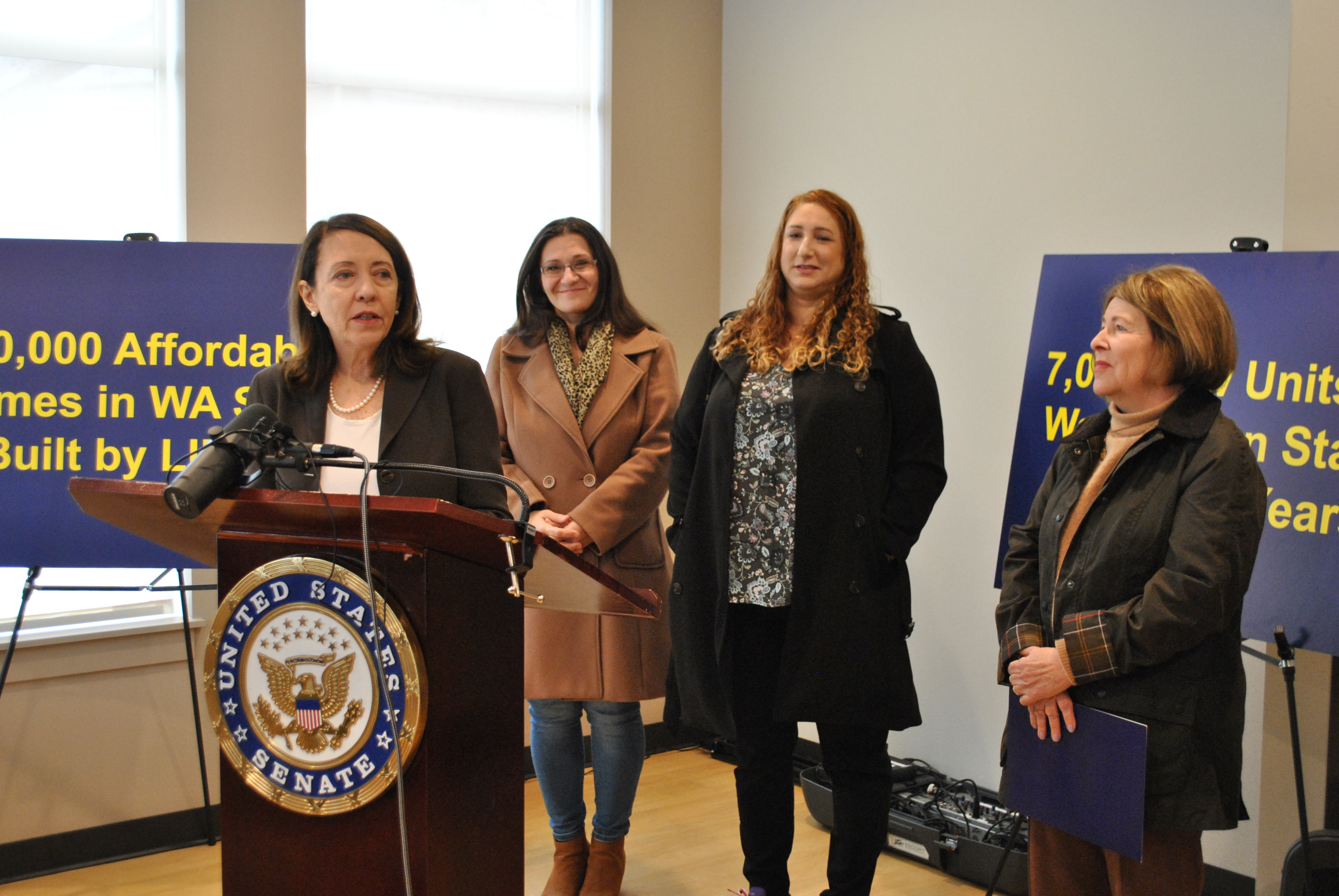 Senator Maria Cantwell stands at a brown podium alongside Spokane Mayor Lisa Brown, while two other affordable housing advocates in winter jackets stand behind them.