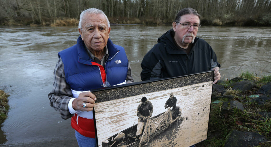 Billy Frank Jr., left, a Nisqually tribal elder who was arrested dozens of times while trying to assert his native fishing rights during the Fish Wars of the 1960s and '70s, poses for a photo Monday, Jan. 13, 2014, with Ed Johnstone, of the Quinault tribe, at Frank's Landing on the Nisqually River in Nisqually, Wash. They are holding a photo from the late 1960s of Frank and Don McCloud fishing on the river. (Credit: Ted S. Warren / AP)