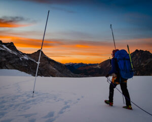 A woman in black snow pants and a yellow jacket with a big blue backpack measures snow with a large rod on top of a mountain covered in snow. An orange sunset is visible in the distance.