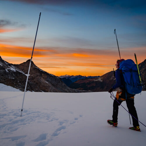 A woman in black snow pants and a yellow jacket with a big blue backpack measures snow with a large rod on top of a mountain covered in snow. An orange sunset is visible in the distance.