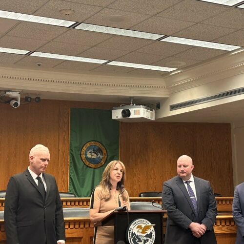 Five people in suits and dresses stand inside a conference room at the Capitol building in Olympia, Washington under fluorescent lights.