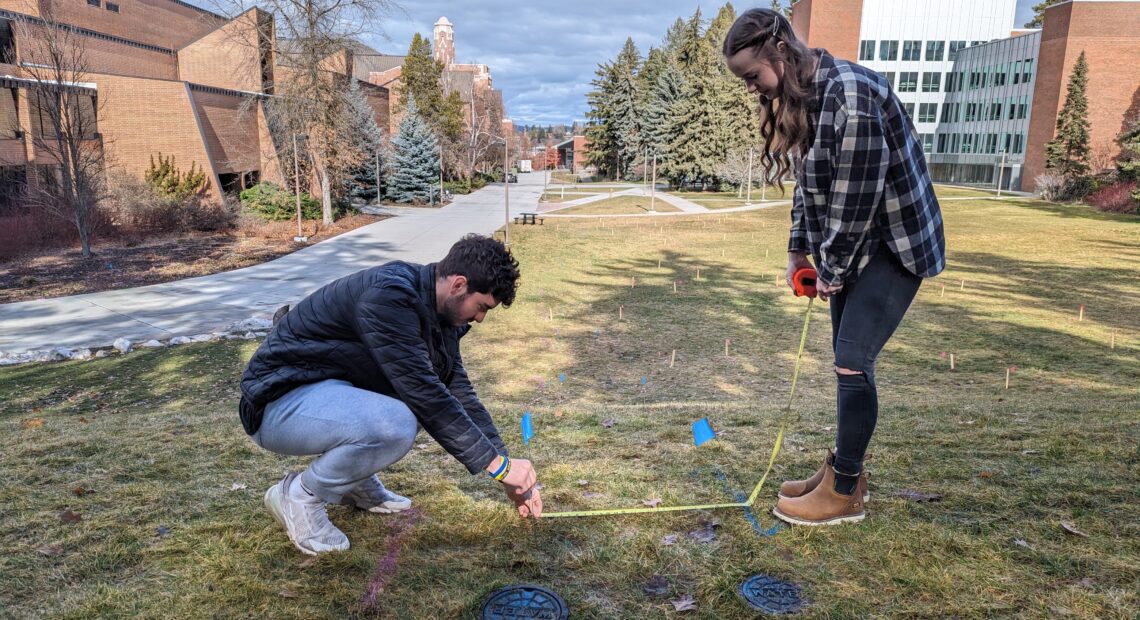 A female student in plaid and jeans and a male student in a black jacket and light jeans use a measuring tape on the ground at the top of a green hill. Brick buildings can be seen in the distance.