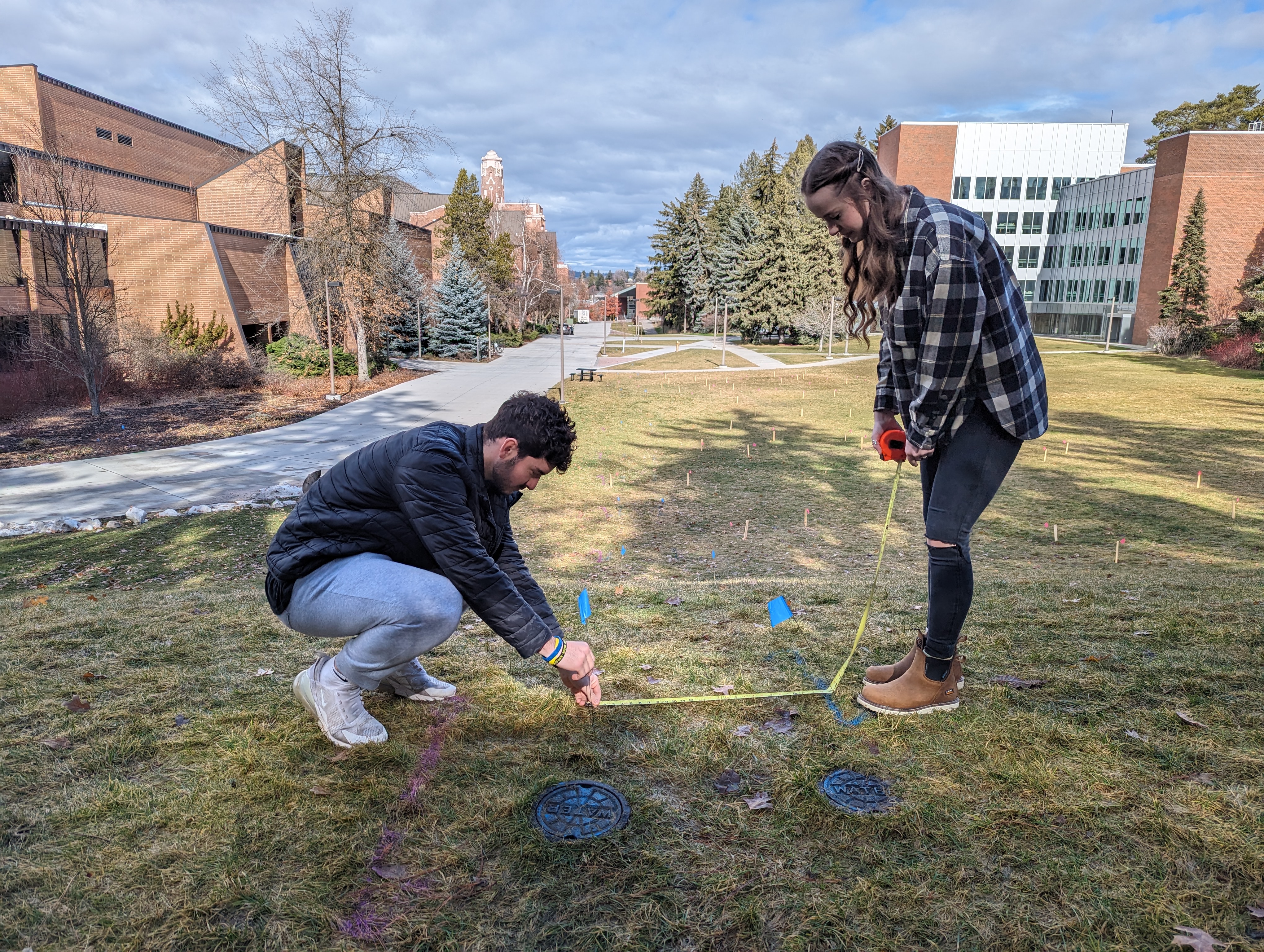 A female student in plaid and jeans and a male student in a black jacket and light jeans use a measuring tape on the ground at the top of a green hill. Brick buildings can be seen in the distance.