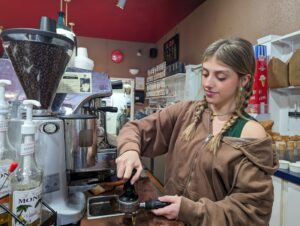 A woman with blonde hair and a brown sweatshirt pulls shots for an Americano at a silver espresso machine.