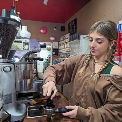 A woman with blonde hair and a brown sweatshirt pulls shots for an Americano at a silver espresso machine.