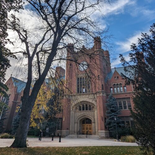 A brick building with a large clock is flanked by evergreen trees under a blue sky.